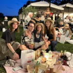 Gruppi di amici seduti sull’erba, circondati da luci decorative, mentre condividono cibo e bevande.