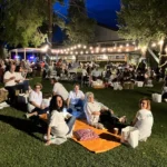 Gruppi di amici seduti sull’erba, circondati da luci decorative, mentre condividono cibo e bevande.