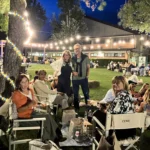 Gruppi di amici seduti sull’erba, circondati da luci decorative, mentre condividono cibo e bevande.