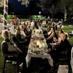 Gruppi di amici seduti sull’erba, circondati da luci decorative, mentre condividono cibo e bevande.
