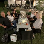 Gruppi di amici seduti sull’erba, circondati da luci decorative, mentre condividono cibo e bevande.