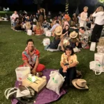 Gruppi di amici seduti sull’erba, circondati da luci decorative, mentre condividono cibo e bevande.