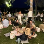Gruppi di amici seduti sull’erba, circondati da luci decorative, mentre condividono cibo e bevande.