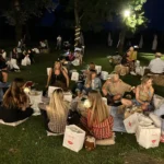 Gruppi di amici seduti sull’erba, circondati da luci decorative, mentre condividono cibo e bevande.