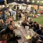 Gruppi di amici seduti sull’erba, circondati da luci decorative, mentre condividono cibo e bevande.