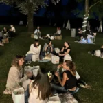 Gruppi di amici seduti sull’erba, circondati da luci decorative, mentre condividono cibo e bevande.
