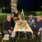 Gruppi di amici seduti sull’erba, circondati da luci decorative, mentre condividono cibo e bevande.
