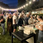 Gruppi di amici seduti sull’erba, circondati da luci decorative, mentre condividono cibo e bevande.