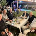 Gruppi di amici seduti sull’erba, circondati da luci decorative, mentre condividono cibo e bevande.