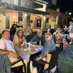 Gruppi di amici seduti sull’erba, circondati da luci decorative, mentre condividono cibo e bevande.