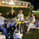 Gruppi di amici seduti sull’erba, circondati da luci decorative, mentre condividono cibo e bevande.