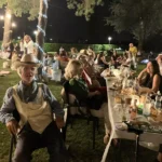 Gruppi di amici seduti sull’erba, circondati da luci decorative, mentre condividono cibo e bevande.