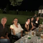 Gruppi di amici seduti sull’erba, circondati da luci decorative, mentre condividono cibo e bevande.